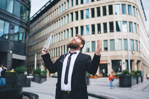 Cheerful businessman in formal suit with tablet and earphones looking up exulting and raising hands while standing on street near modern buildings - Valokuva, kuva