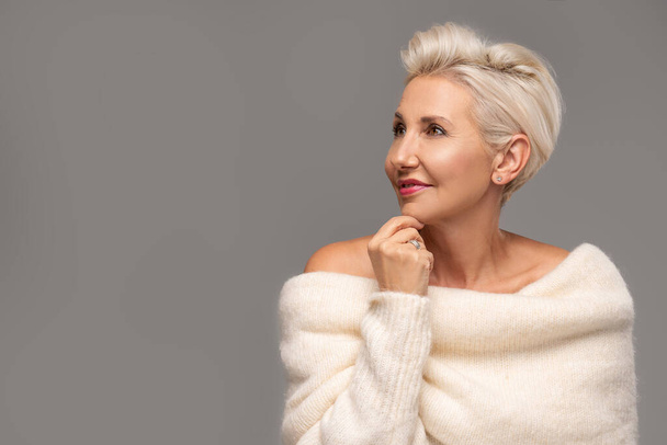 Beauty portrait of elegant adult blonde woman with short hairstyle and glamour makeup. Attractive lady wearing warm fashionable sweater. A lot of copy space. Studio shot. - 写真・画像
