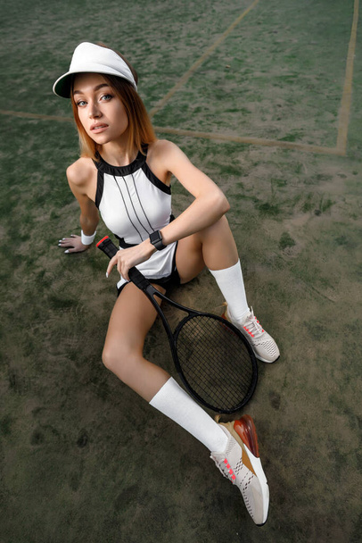 a young blond woman with a tennis racket in white clothes and a cap sits on a tennis court. sunny - 写真・画像