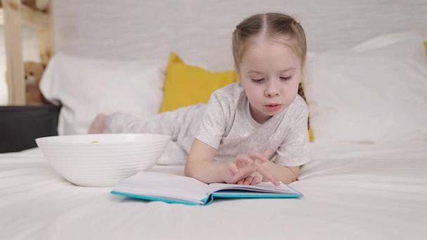 little child eats reading book while lying bed, happy family, kid daughter doing homework and eating potato chips, doing homework elementary school, first grader loves literature, children school life - Фото, изображение