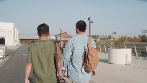 Tracking back-view medium shot van twee jonge Aziatische mannen in casualwear wandelen buiten in de zomer langs dijk - Video