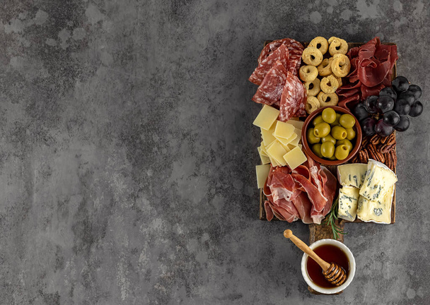 Вид сверху вкусной картонной доски с сыром, виноградом, орехами, оливками и ветчиной на круглой кухонной тарелке - Фото, изображение