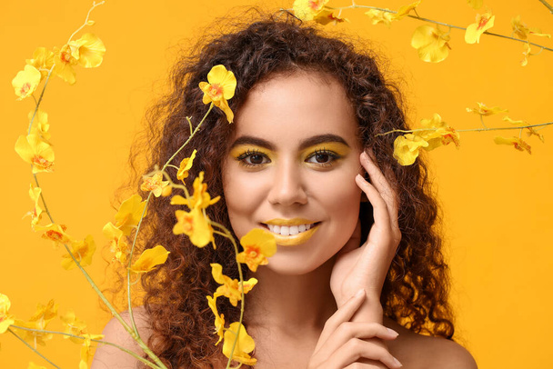 Beautiful African-American woman with makeup in yellow tones and flowers on color background - Photo, Image