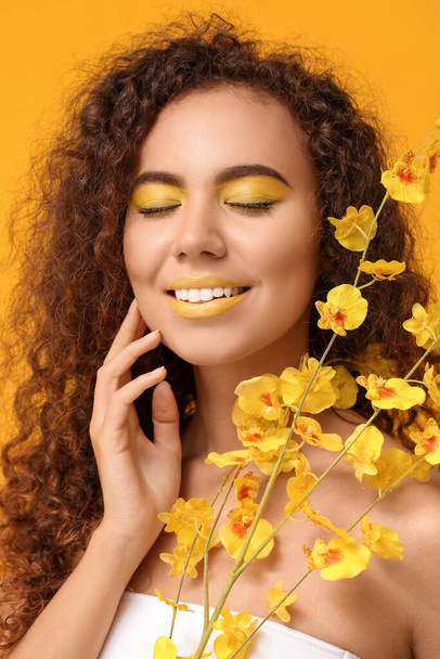 Hermosa mujer afroamericana con maquillaje en tonos amarillos y flores sobre fondo de color - Foto, Imagen