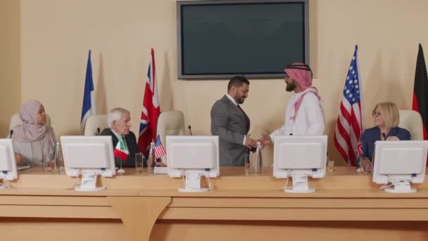 Stab shot of two foreign male politicians shaking hands at official press conference with representatives of different countries, sitting at long table equipped with computer monitors and microphones - Filmati, video