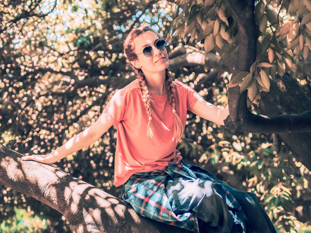 Dame aux lunettes de soleil avec deux nattes et une chemise attachée sur une ceinture se trouve sur un arbre dans un parc d'été. Concentration floue - Photo, image