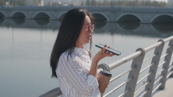 Medium shot of young Asian woman in sunglasses recording voice message on cellphone standing at river embankment on summer day - Filmati, video