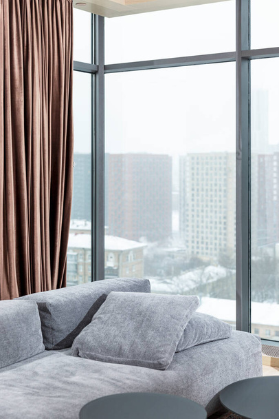 The interior of a stylish living room with panoramic windows in a city high-rise building. Gray cozy sofa and brown curtains. Vertical. - 写真・画像