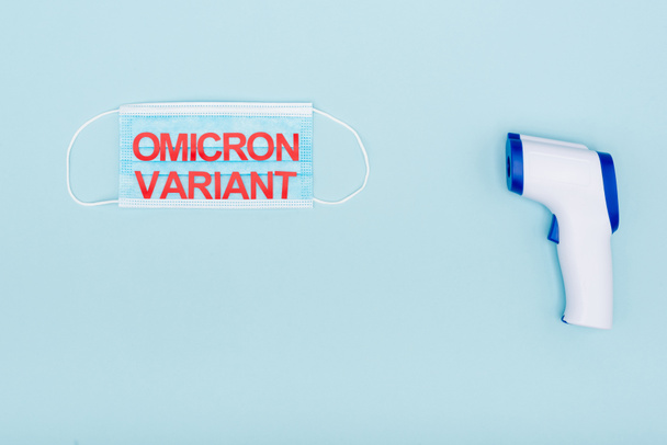 top view of medical mask with red omicron variant lettering near pyrometer on blue - Foto, Imagem