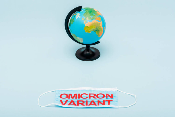 medical mask with red omicron variant lettering near globe on blue background - Fotografie, Obrázek