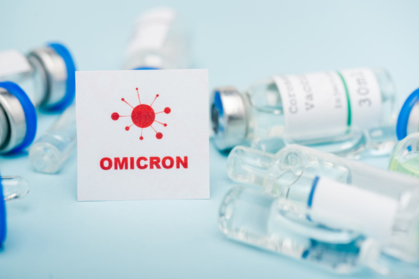 white card with red omicron lettering and bacteria near blurred vaccine vials and ampoules on blue - Foto, Imagen