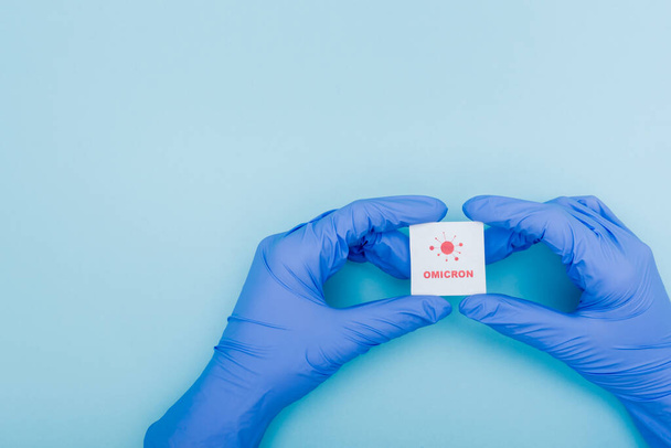 white card with red omicron lettering and bacteria icon in hands of doctor in latex gloves on blue, cropped view - Foto, Imagem