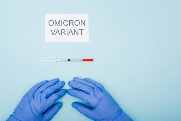 top view of doctor in latex gloves near card with omicron variant lettering and syringe on blue - Foto, Imagem
