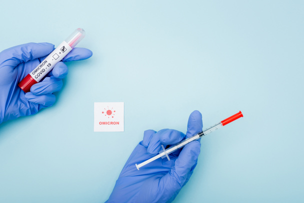 top view of doctor in latex gloves holding syringe and test tube near card with omicron lettering and bacteria on blue - Foto, afbeelding