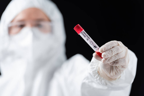 científico borroso en traje de hazmat blanco con prueba positiva de variante omicrónica covid-19 aislada en negro - Foto, Imagen