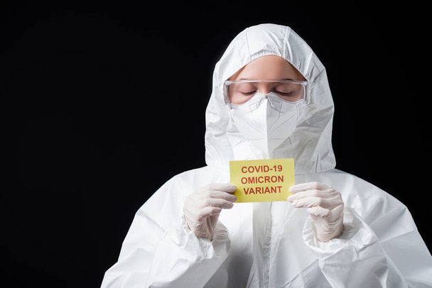 scientist in white hazmat suit and goggles holding card with covid-19 omicron variant lettering isolated on black - Photo, Image