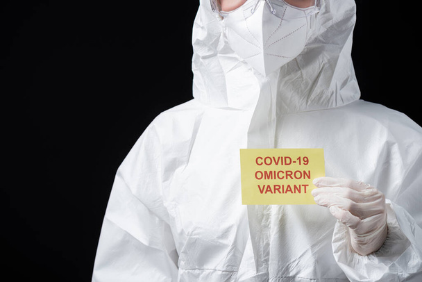 vista parcial del médico en la tarjeta de retención de traje de materiales peligrosos con letras variantes omicrónicas covid-19 aisladas en negro - Foto, imagen