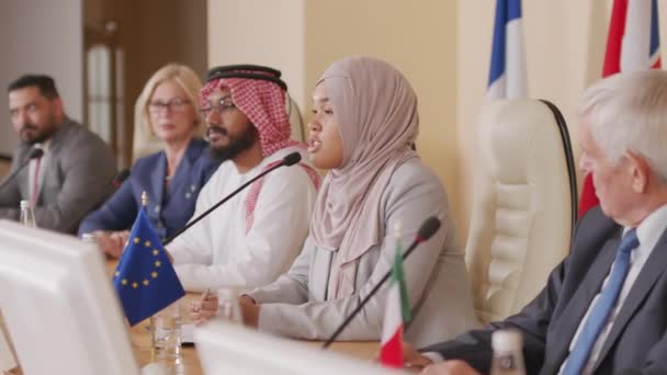 Střední záběr mladé muslimky politického lídra v hidžábu, jak pronáší projev v mikrofonu na oficiální tiskové konferenci v interiéru, sedí u stolu se zástupci různých zemí - Záběry, video