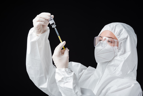 immunologist in personal protective equipment holding syringe and covid-19 omicron variant vaccine isolated on black - Foto, immagini