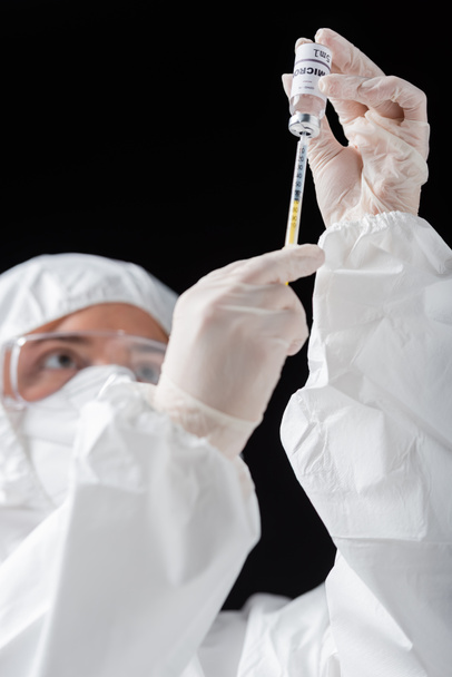 blurred doctor in personal protective equipment holding syringe and omicron variant vaccine isolated on black - Фото, зображення