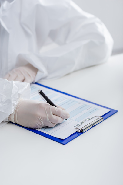 partial view of blurred doctor in hazmat suit writing in medical card on clipboard in lab - Foto, Imagem