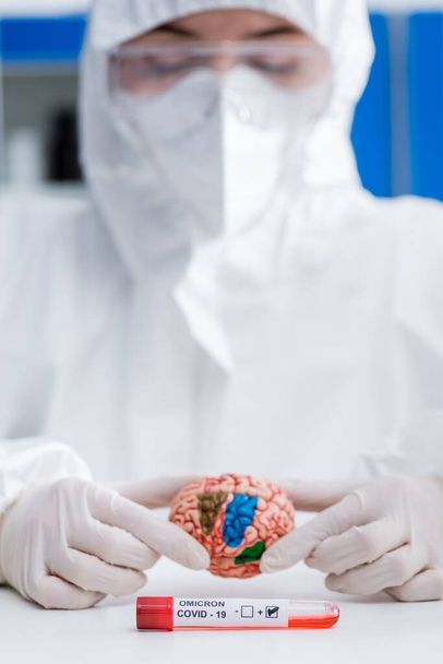 médico borroso en traje de materiales peligrosos que sostiene el modelo cerebral cerca de la prueba de variante de omicrón covid-19 positivo - Foto, Imagen