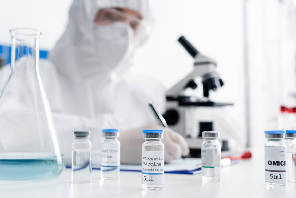 selective focus of vials with coronavirus vaccine near scientist writing near microscope on blurred background - Фото, изображение