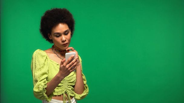 mujer afroamericana pensativa usando teléfono inteligente aislado en verde  - Foto, imagen