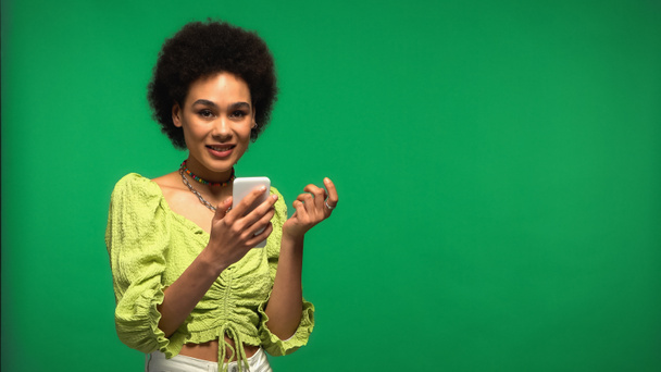 mujer afroamericana feliz usando teléfono inteligente aislado en verde  - Foto, imagen