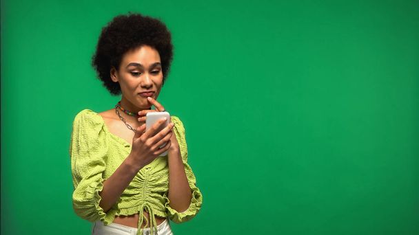 mujer afroamericana reflexiva usando teléfono inteligente aislado en verde  - Foto, Imagen