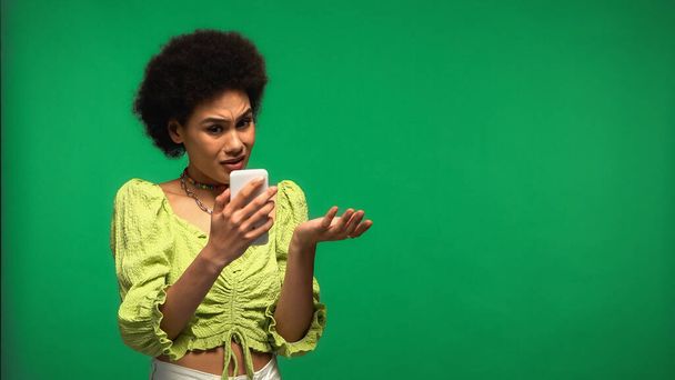 nespokojený africký Američan žena při pohledu na smartphone izolované na zelené  - Fotografie, Obrázek