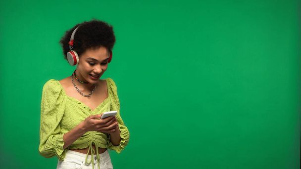 šťastný africký Američan žena v bezdrátových sluchátek pomocí smartphone izolované na zelené  - Fotografie, Obrázek