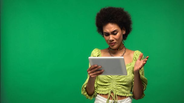 mujer afroamericana disgustada en blusa usando tableta digital aislada en verde  - Foto, Imagen