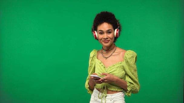 alegre mujer afroamericana en blusa y auriculares inalámbricos usando teléfono inteligente aislado en verde  - Foto, Imagen