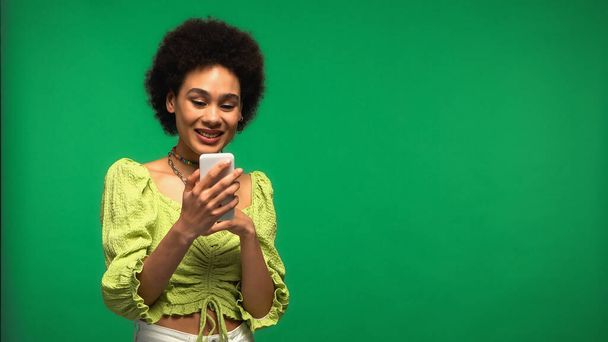 mujer afroamericana feliz en blusa mirando teléfono inteligente aislado en verde  - Foto, imagen