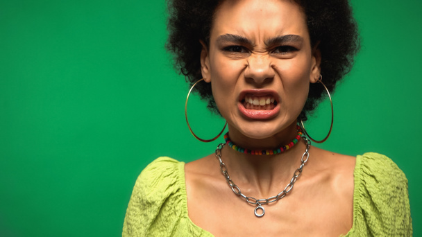 mujer afroamericana irritada en pendientes de aro mirando a la cámara aislada en verde  - Foto, Imagen