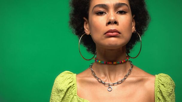dissatisfied african american woman in hoop earrings looking at camera isolated on green - 写真・画像