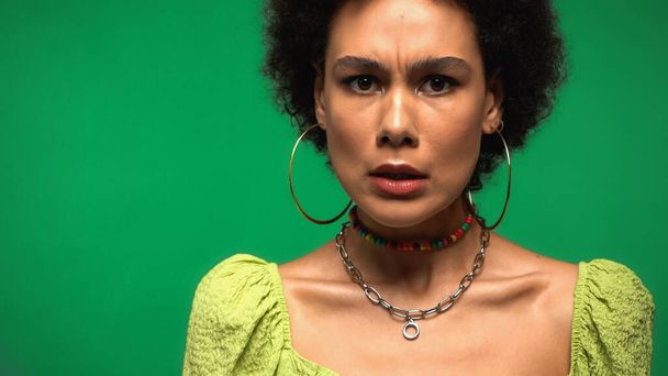 curly african american woman in hoop earrings grimacing isolated on green - Photo, Image