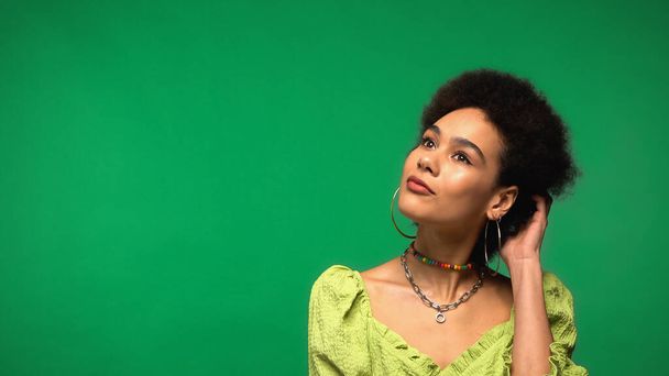 dreamy african american woman in hoop earrings adjusting curly hair isolated on green - 写真・画像