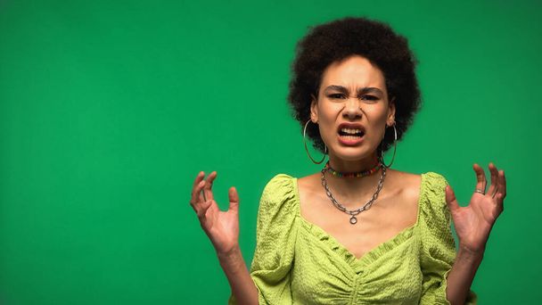 geïrriteerde Afro-Amerikaanse vrouw in hoepel oorbellen gebaren en kijken naar camera geïsoleerd op groen  - Foto, afbeelding