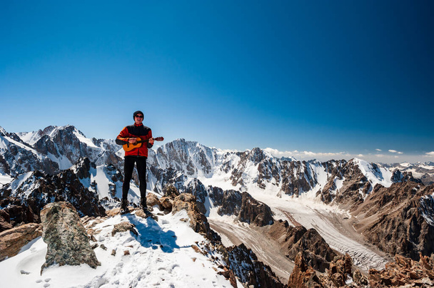 Młody człowiek trekker gra na gitarze i śpiewa piosenkę na szczycie Pik Uchitel szczyt. Krajobraz alpejskiego parku narodowego Ala Archa w pobliżu Biszkek, pasmo górskie Tian Shan, Kirgistan, Azja Środkowa. - Zdjęcie, obraz