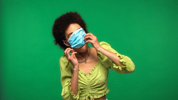 curly african american woman wearing blue medical mask isolated on green - Photo, Image