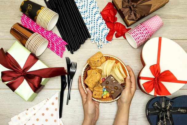 щаслива концепція дня народження, жінка тримає шоколадну цукерку тарілку та соломинку та чіпси картоплю, печиво та одноразові чашки з пластиковими виделками та подарунковими коробками на дерев'яному столі
 - Фото, зображення