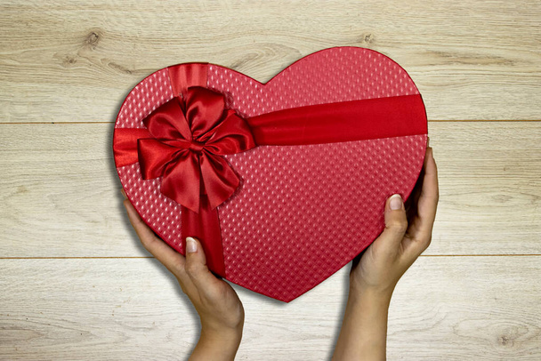 χέρια γυναίκα κρατώντας κόκκινο κουτί σχήμα καρδιάς απομονώνονται σε ξύλινο τραπέζι, έννοια διακοπές - Φωτογραφία, εικόνα