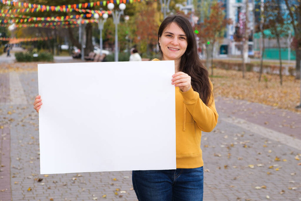 Μια μελαχρινή γυναίκα χαμογελά και κρατά ένα άδειο λευκό φύλλο για την επιγραφή και το κείμενο - Φωτογραφία, εικόνα