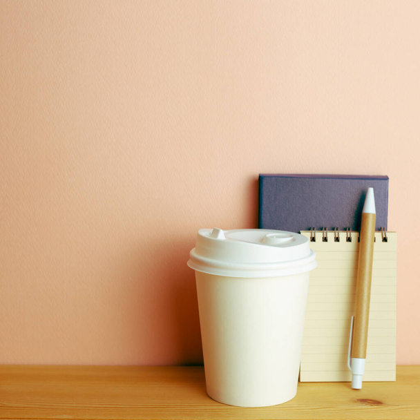 Notatnik, długopis, papierowy kubek na drewnianym biurku. różowy tło ściany. przestrzeń robocza - Zdjęcie, obraz
