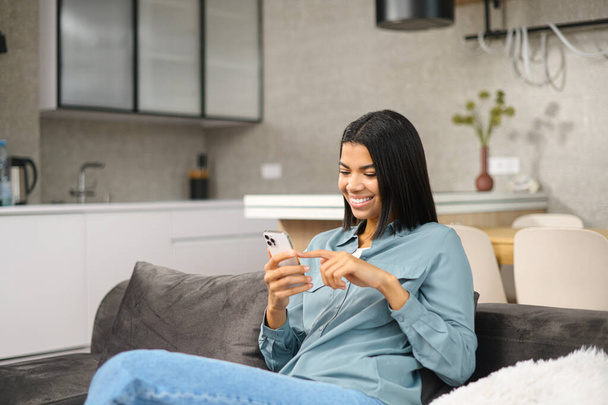 Glad brunette woman spending leisure with a smartphone, checking emails, shopping online, scrolling news feed sitting on the comfortable couch at home. Smiling female web surfing - Fotoğraf, Görsel
