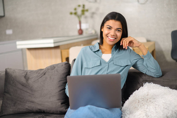 Young mixed-race female freelancer with brunette hair in comfy casual clothes sitting on the couch and working on the project, typing email on the laptop - 写真・画像