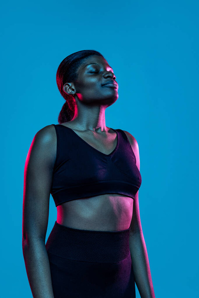 Portrait of young beautiful dark skinned woman posing isolated on blue studio background in neon light. - Foto, Bild