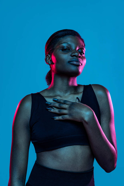 Portrait of young beautiful dark skinned woman posing isolated on blue studio background in neon light. - Fotoğraf, Görsel